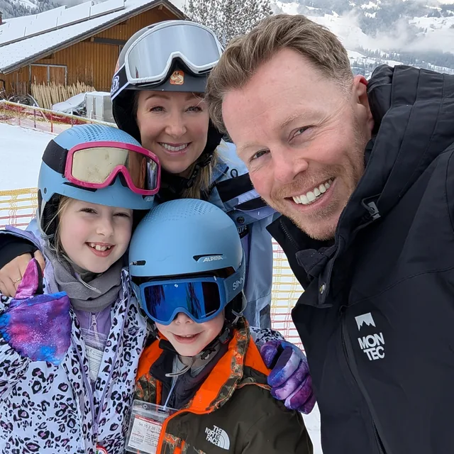 Family selfie on the slopes: Matthew in a black ski jacket with his wife and two kids in helmets and goggles, snowy chalets and mountains behind.