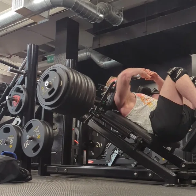 Matthew on a loaded leg press in an empty gym, shot low from the rubber floor, plates stacked on the rack.
