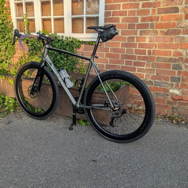 A grey road bike with deep-section carbon wheels leaning against an ivy-covered brick wall in evening sun.